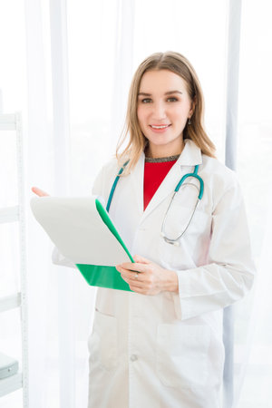 A female beautiful doctor is wearing a doctor uniform have a stethoscope and holding a green paper chart, smiling and looking at the camera the background is a white curtain in the hospital or clinic.の写真素材
