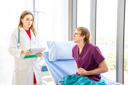 The male patient, wearing in a patient's dress, had abdominal pain, sitting on a patient bed by a window in a hospital or clinic room with a window background and white curtains.の写真素材