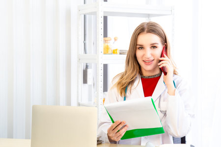 A female beautiful doctor is wearing a doctor uniform have a stethoscope and holding a green paper chart and talking on mobile phone in the examination room in the hospital or clinic.の写真素材