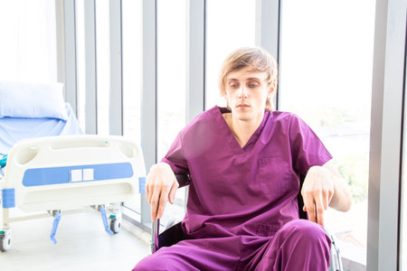 The male patient wearing in a patient dress was disheartened or hopeless, sitting on a wheelchair in the patient room background by a window in a hospital or clinic room.の写真素材