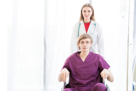 The male patient wearing in a patient dress in a wheelchair and female beautiful doctor is wearing a doctor uniform have a stethoscope and holding a green paper standing on the side.の写真素材