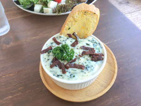 Baked spinach with cheese and topped with crispy bacon served with butter bread in a white ceramic bowl on a wooden tray on a wood table in the restaurant.の写真素材