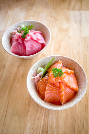 Salmon don and Maguro or tuna don Consisting of sliced salmon and Maguro or tuna on top of Japanese rice Served with wasabi and pickled ginger in a white bowl on a wooden table.の写真素材