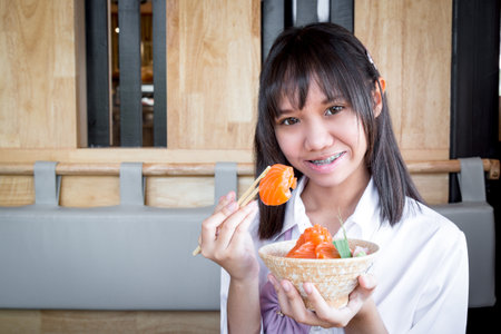 A girl teenage ethnic Asian shows a raw salmon uses chopsticks to pick up from salmon don with rice and smiling in a Japanese restaurant. Selective focus.の写真素材
