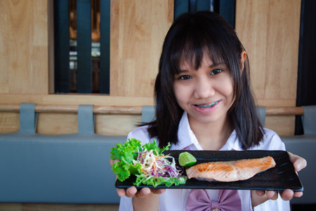 A girl teenage shows a Salted Grilled Salmon served with lemon and salad to the front and looking at the camera and smiling in a Japanese restaurant with copy space. Selective focus.の写真素材