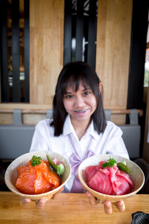 A girl teenage shows a salmon don and maguro or tuna don is a raw salmon and maguro or tuna with rice to the front and looking at the camera and smiling in a Japanese restaurant. Selective focus.の写真素材
