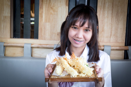 A girl teenage shows a Shrimp Tempura on bamboo plate to the front and looking at the camera and smiling in a Japanese restaurant with copy space. Selective focus.の写真素材