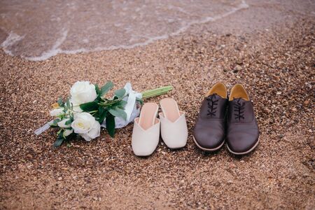 Close-up photos of a bouquet of flowers and shoes, the groom and the bride, placed on the sandy beach with ocean waves as the evening sunset background.の写真素材