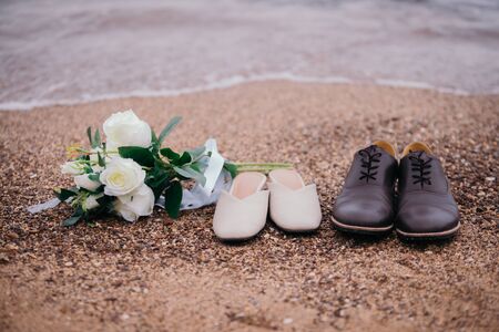 Close-up photos of a bouquet of flowers and shoes, the groom and the bride, placed on the sandy beach with ocean waves as the evening sunset background.の写真素材