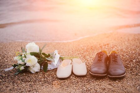 Close-up photos of a bouquet of flowers and shoes, the groom and the bride, placed on the sandy beach with ocean waves as the evening sunset background.の写真素材
