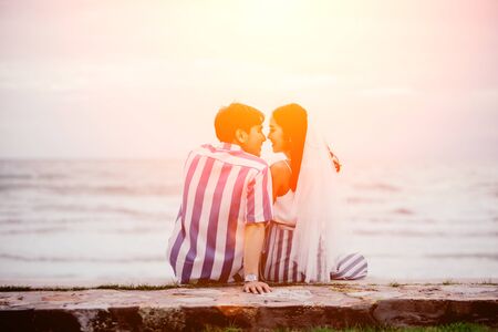 Rear view of a happy couple sitting embrace and lean on the shoulder the beach watching the sunset in summer. Men and women with love are showing through the body by embracing.の写真素材