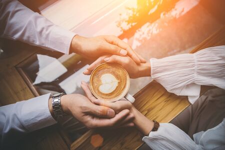 Valentine day, Top view of a young lovers hand holding a heart shaped coffee cup on a wooden table in the coffee shop. Warm Tone.の写真素材