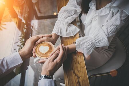 Valentine day, Close up of a young lovers hand holding a heart shaped coffee cup on a wooden table in the coffee shop. Warm Tone. Couple wedding.の写真素材