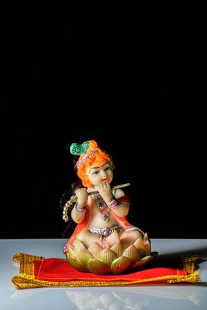 An isolated sculpture of Indian god lord Krishna playing flute sitting on a mat. On a reflective white table with a dark background.Portrait viewの写真素材