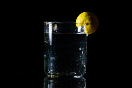 A glass of water placed on a reflective surface in a dark background and a cut lemon on the edge of the glassの写真素材