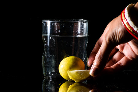 A glass of water placed on a reflective surface in a dark background with cut and intact lemon...