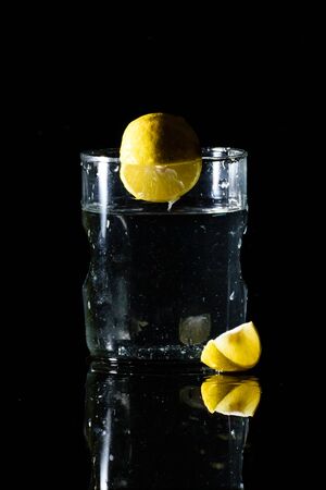 A glass of water placed on a reflective surface in a dark background with a cut lemon placed in front of it and an intact lemon on the edge of the glassの写真素材