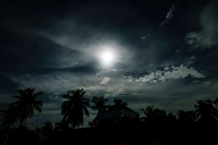 A full moon night in a village with clouds above and buildings and trees below with halo surrounding the moonの写真素材