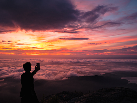 Silhouette People Take a Selfie on the cliff with beautiful sunrise sky on Khao Luang mountain in Ramkhamhaeng National Park,Sukhothai province Thailandの写真素材