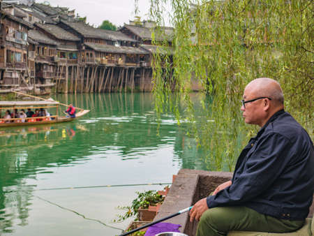 fenghuang,Hunan/China-16 October 2018:Unacquainted Chinese old man fishing in the river of fenghuang old town .phoenix ancient town or Fenghuang County is a county of Hunan Province, Chinaのeditorial素材