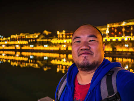 Portrait Photo of Asian fat backpacker with beautiful Scenery view in the night of fenghuang old town .phoenix ancient town or Fenghuang County is a county of Hunan Province, Chinaの写真素材