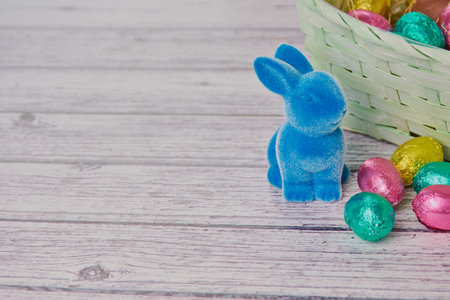 blue easter bunny beside colored chocolate eggs, in the background an easter basket on white wooden...