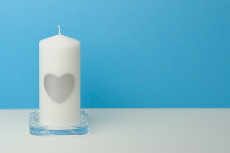 white big candle with a silver heart on it in front of a blue background on a white table, with copy space for greeting cardsの写真素材