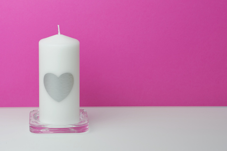 white big candle with a silver heart on it in front of a pink background on a white table, with copy space, for greeting cardsの写真素材