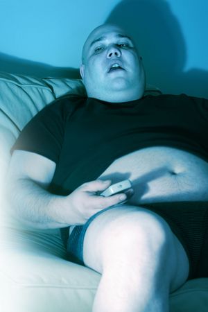 Lazy overweight male sitting on a couch watching television.  Harsh blue lighting from television with slow shutter speed to create TV watching atmosphere. Selective focus on the eyes.の写真素材