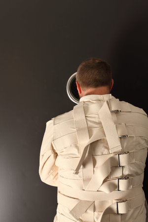 Photo of a insane man in his forties wearing a straitjacket looking out the hole of an asylum door.