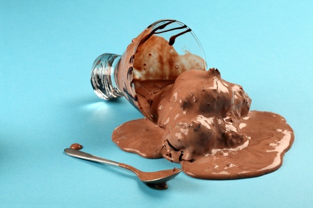 Photo of a bowl of melting chocolate ice cream tipped over and melting across a blue table.の写真素材