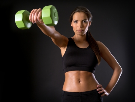 Photo of a beautiful female doing a front shoulder fly with a dumbbell over a dark background.の写真素材