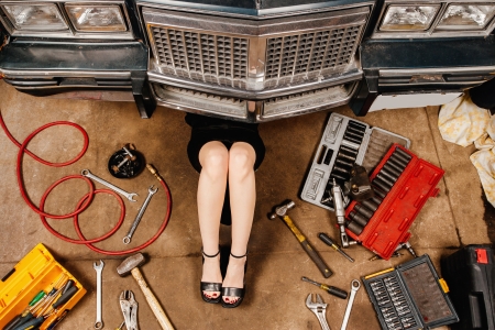 A woman wearing a black skirt and heels doing repairs under the front of an old car from the early 80's.の写真素材