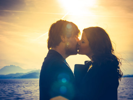 Photo of young adults by the water hugging and kissing in the late afternoon as the sun lowers in...