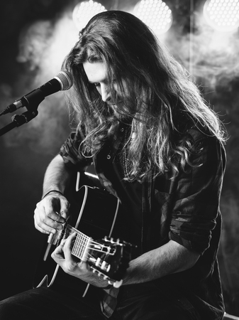 Photo of a young man with long hair and a beard singing and playing an acoustic guitar on stage with lights and concert atmosphere.の写真素材