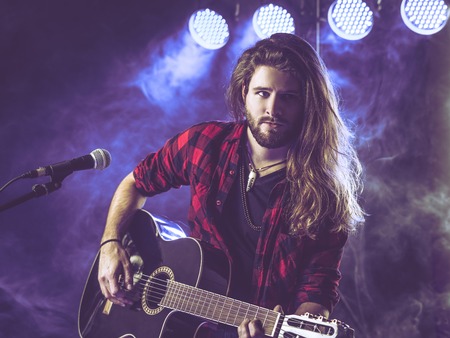 Photo of a young man with long hair and a beard playing an acoustic guitar on stage with lights and concert atmosphere.の写真素材