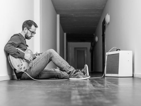 Photo of a man sitting playing his electric guitar in front of a small combo amplifier in a hallway....