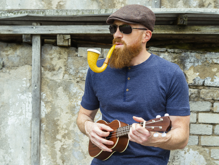 Photo of a bearded man in his thirties smoking a large pipe and playing a ukulele.の写真素材