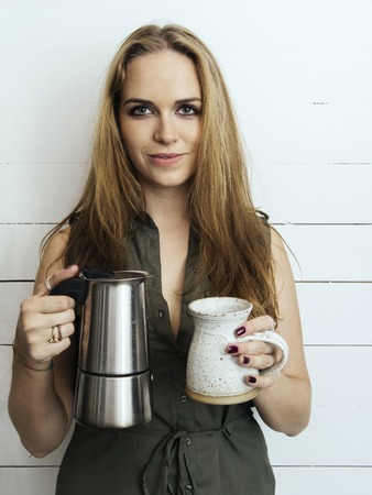 Photo of a beautiful smiling woman holding a Moka pot and coffee mug and looking at the camera.の写真素材