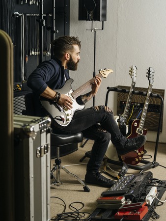 Photo of a man with beard sitting and playing his electric guitar in a recording studio.の写真素材