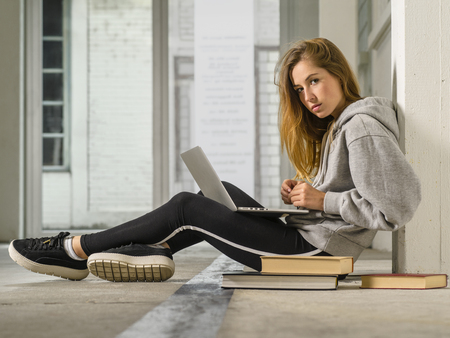 Photo of a young woman student sitting in a hallway with a laptop and books.の写真素材