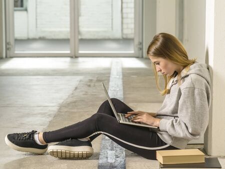 Photo of a young woman student sitting in a hallway with a laptop and books.の写真素材