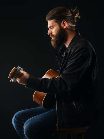 Young bearded man sitting and playing an acoustic guitar over black background.の写真素材