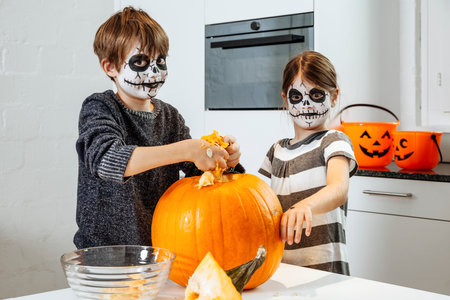 Two young children with skull face paint carving a pumpkin in their kitchen.の写真素材