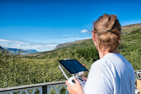 Young man flying a drone in a rural setting on bright summer day, holding the remote control and...