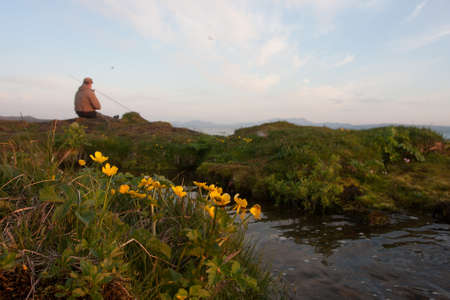Fisherman sitting on the bank of a mountain river with yellow flowersの写真素材