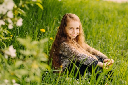 Little cute fashionable long-haired brown-eyed girl sitting on the lawn in blossoming spring garden outdoorの写真素材