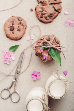 Pile of chocolate chip cookies with two bottles of milk, pink flowers and scissors on rustic grey...
