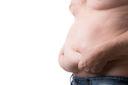 Guy shows with his hands his fat folds on his belly closeup. Overweight caucasian man in blue jeans pinches his tummy on white background. Obesity and excess weight concept. Copy space at the leftの写真素材