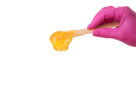 Female hand in pink medical gloves holding wooden spatula with sugaring paste for depilation procedure isolated on white background, hair removal concept, copy spaceの写真素材